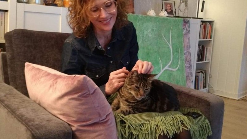 Kvinne sitter i sofa i en stue med et pledd og en katt på fanget