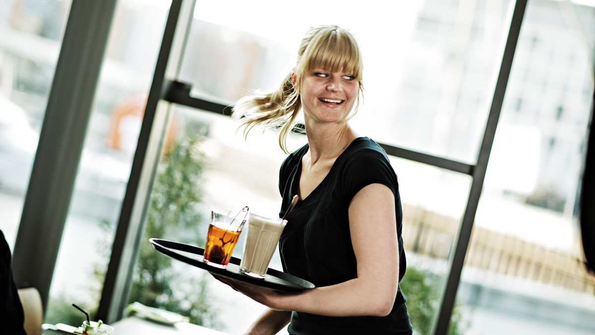 Kvinnelig servitør som ser travel men smilende ut og holder et brett med en kaffe og en kopp te. 