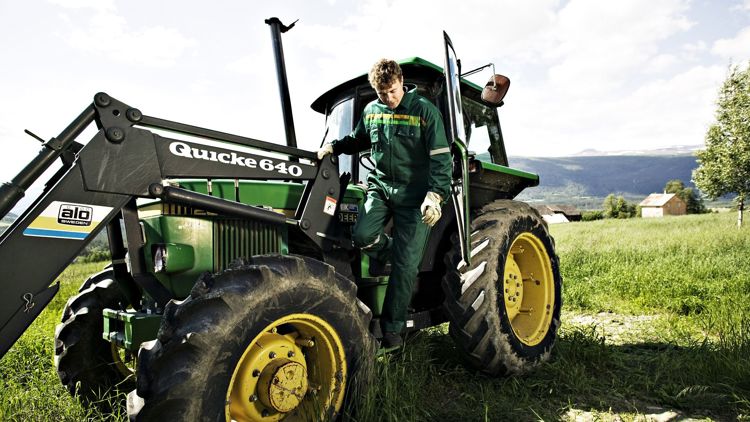 Fotografi av en mann i grønn kjeledress som stiger ut av en traktor på et jorde.