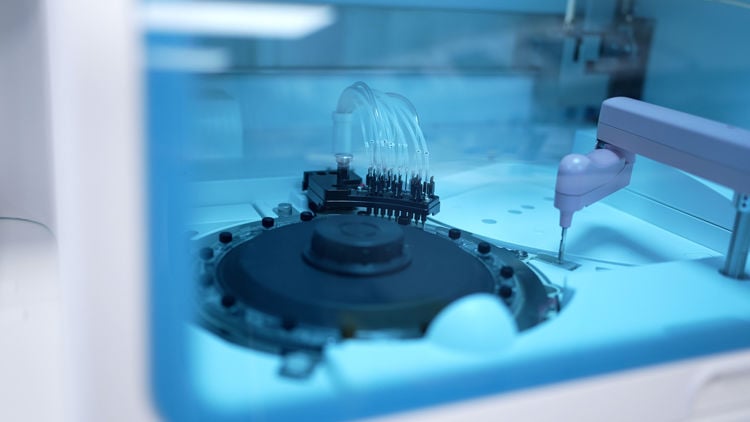 Nærbilde av en automatisert laboratorie- eller industrimaskin med robotarm og prøvehåndteringsutstyr.