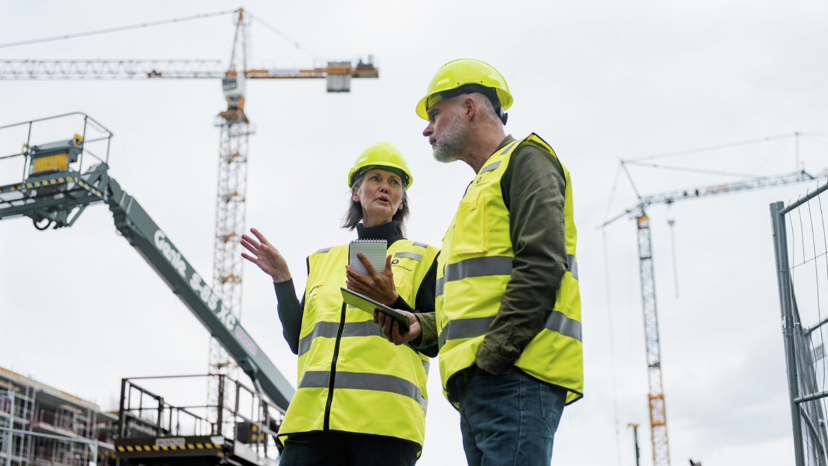 Mann og kvinne med hjelm og refleksvest står og snakker på en byggeplass med to heisekraner i bakgrunnen