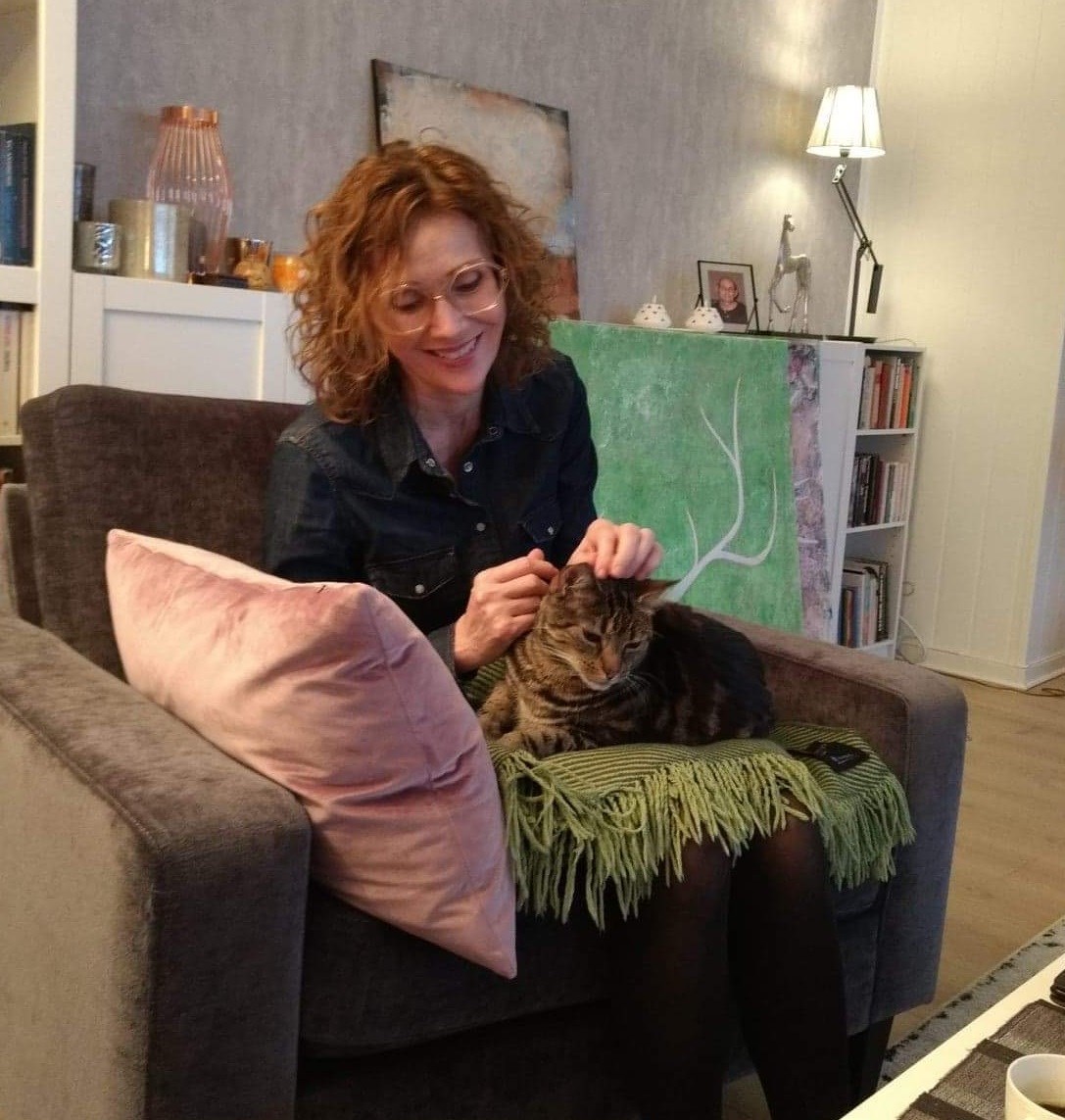 Kvinne sitter i sofa i en stue med et pledd og en katt på fanget