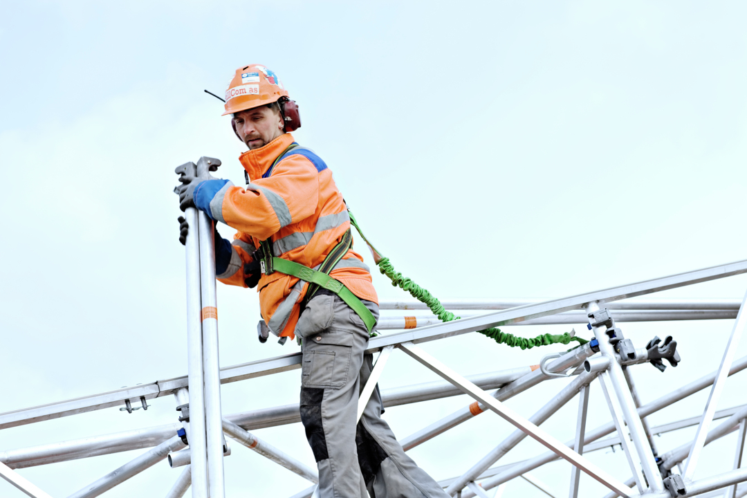 Mann med hjelm og sikkerhetsstrikk festet til rekkverk jobber i høyden 