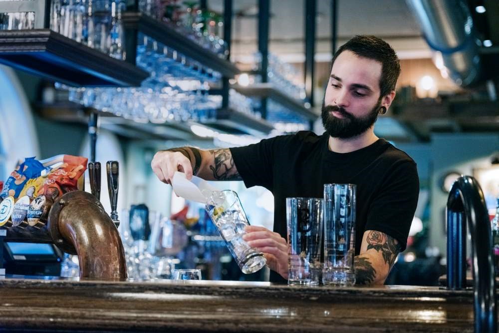 Bildet viser en bartender som står bak disken og lager en drink. 