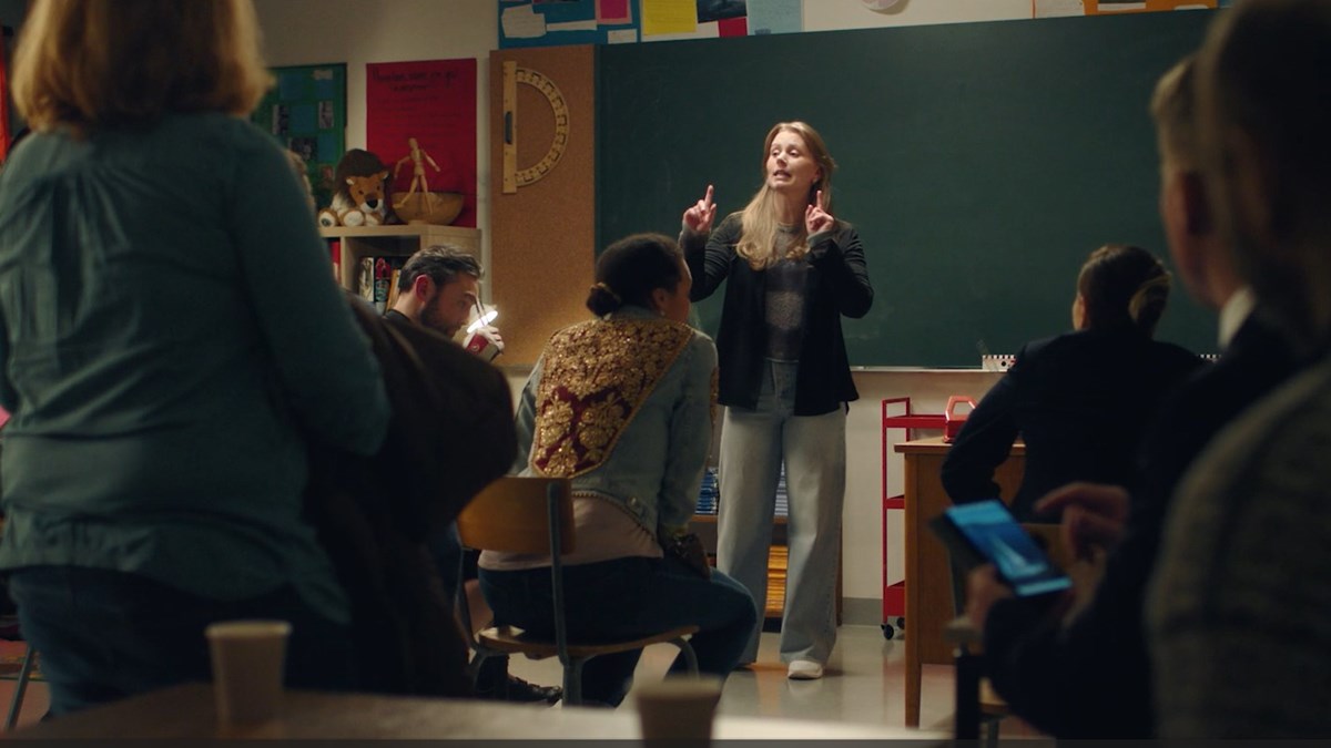 Bildet viser en lærer som står foran ei tavle. Man ser ryggen på flere voksne som sitter og står i klasserommet. 