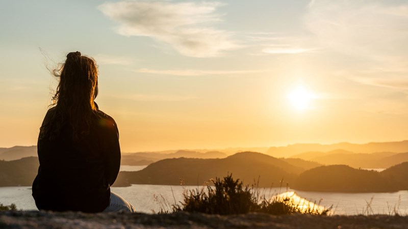 Bildet viser ryggen på ei kvinne som sitter på toppen av et fjell og ser på havet og solnedgangen. 