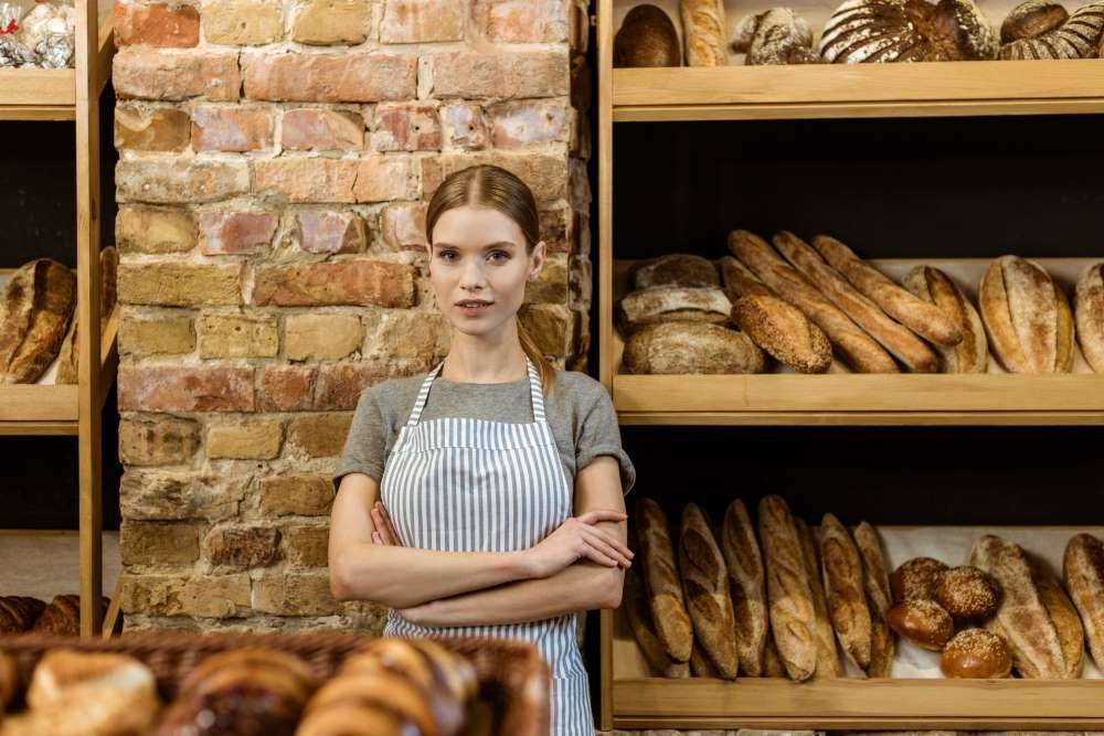 Bildet viser en ung kvinne som jobber på et bakeri og som står foran ei hylle med bakevarer. 