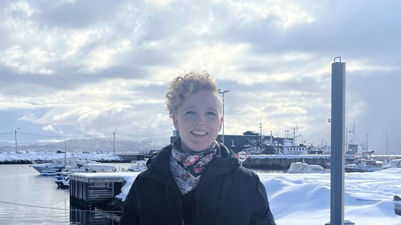 Bilde av inspektør Anne Orheim, smilende  foran ei småbåthavn, med en fabrikkbygning i bakgrunnen