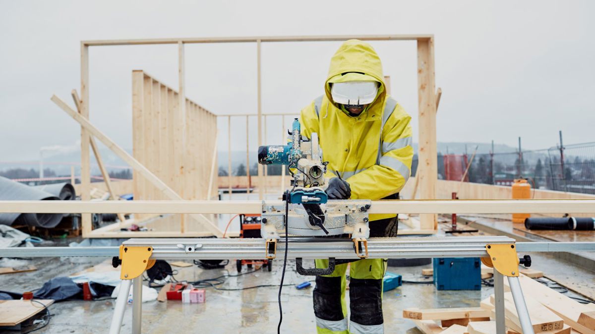 BIldet viser en mann som står på en utendørs byggeplass og jobber ved ei sag.