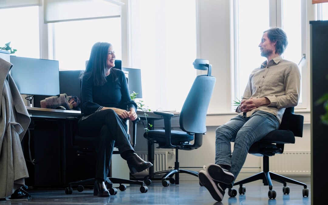 Bildet viser en mann og ei kvinne som sitter i et åpent kontorlandskap. De snakker sammen. 