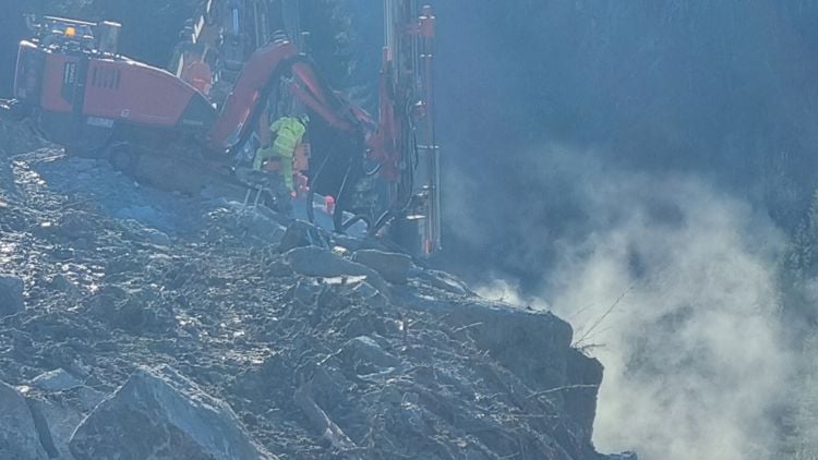 En borerigg i arbeid i et steinbrudd med en sky av steinstøv i bakgrunnen