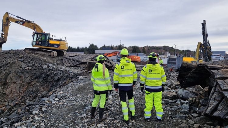 Tre personer står i gule arbeidsklær i et grustak med maskiner i nærheten 