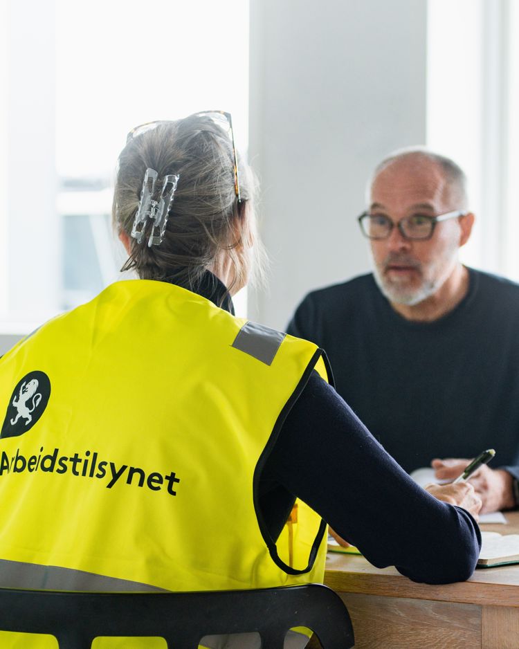 en kvinnelig inspektør fra Arbeidstilsynet i gul vest og en mann sitter ved et bord og snakker sammen